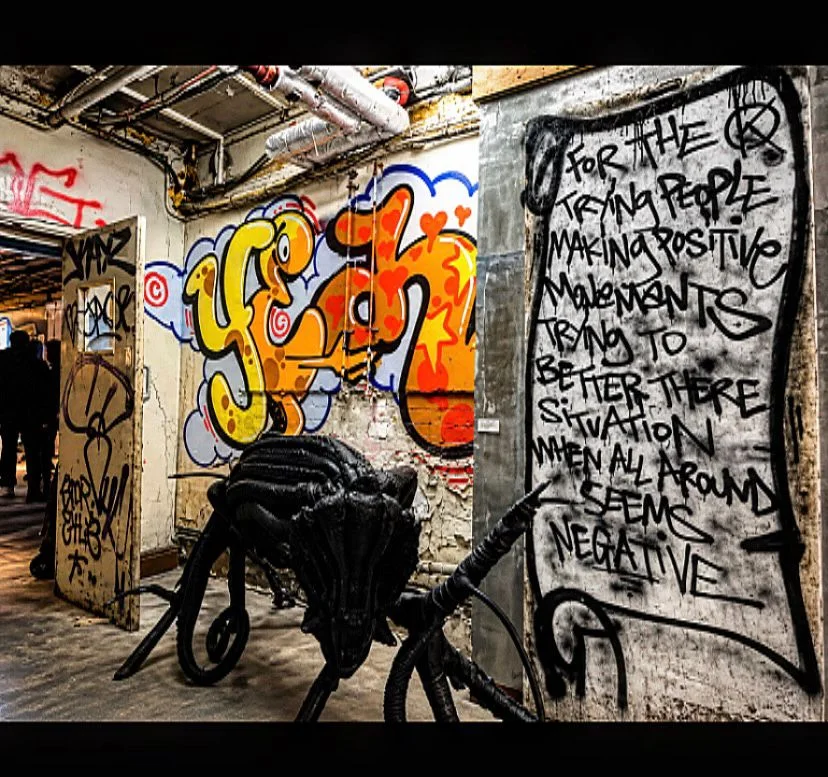 Industrial art space with colourful graffiti lettering, handwritten wall text and a dark sculptural creature