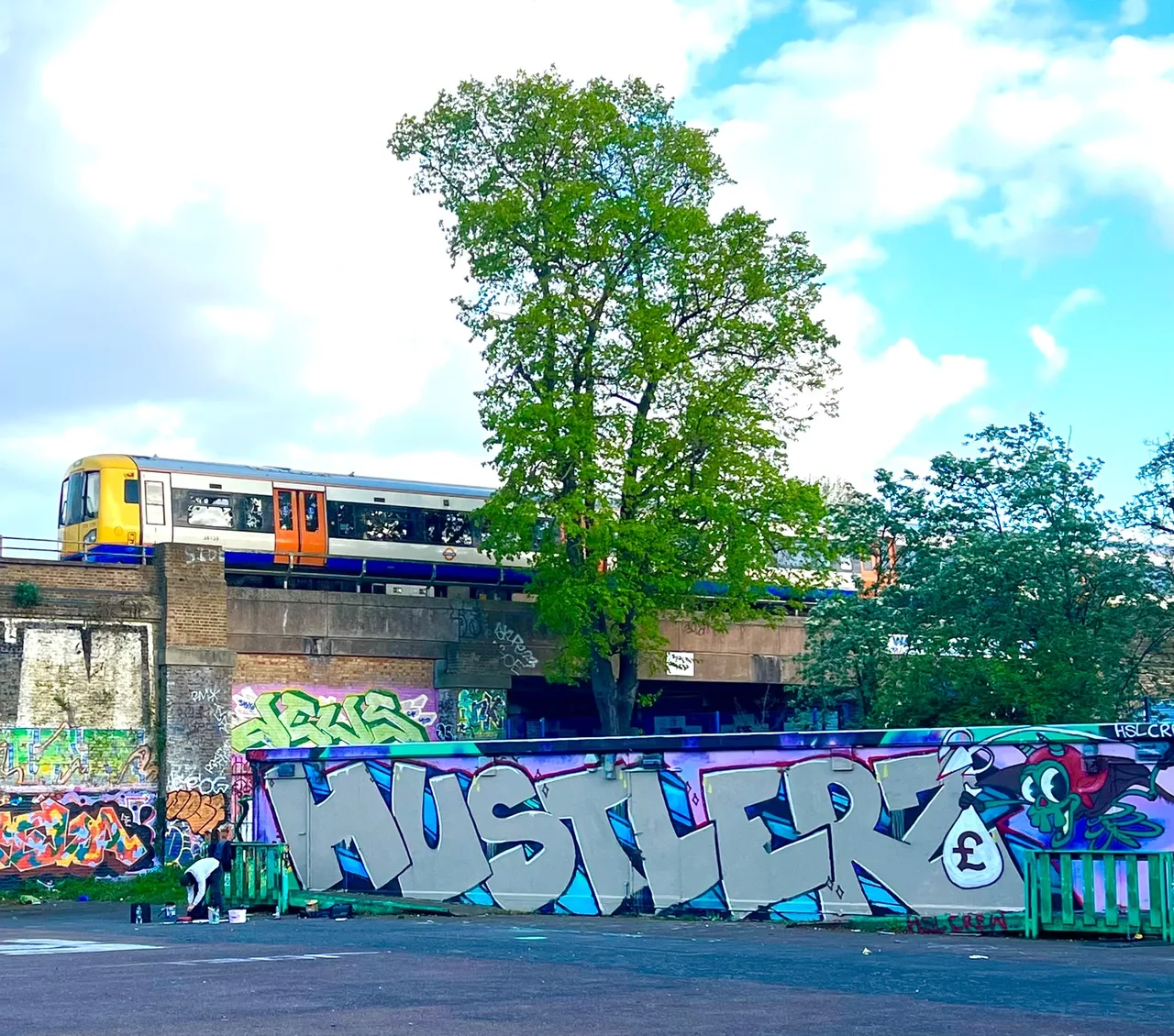 Silver graffiti lettering across an outdoor wall beside a railway, with a train passing above